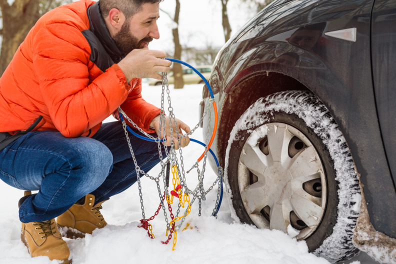 A man in an orange puffer jacket crouching in the snow next to a car tire, holding and untangling a set of metal tire chains.
