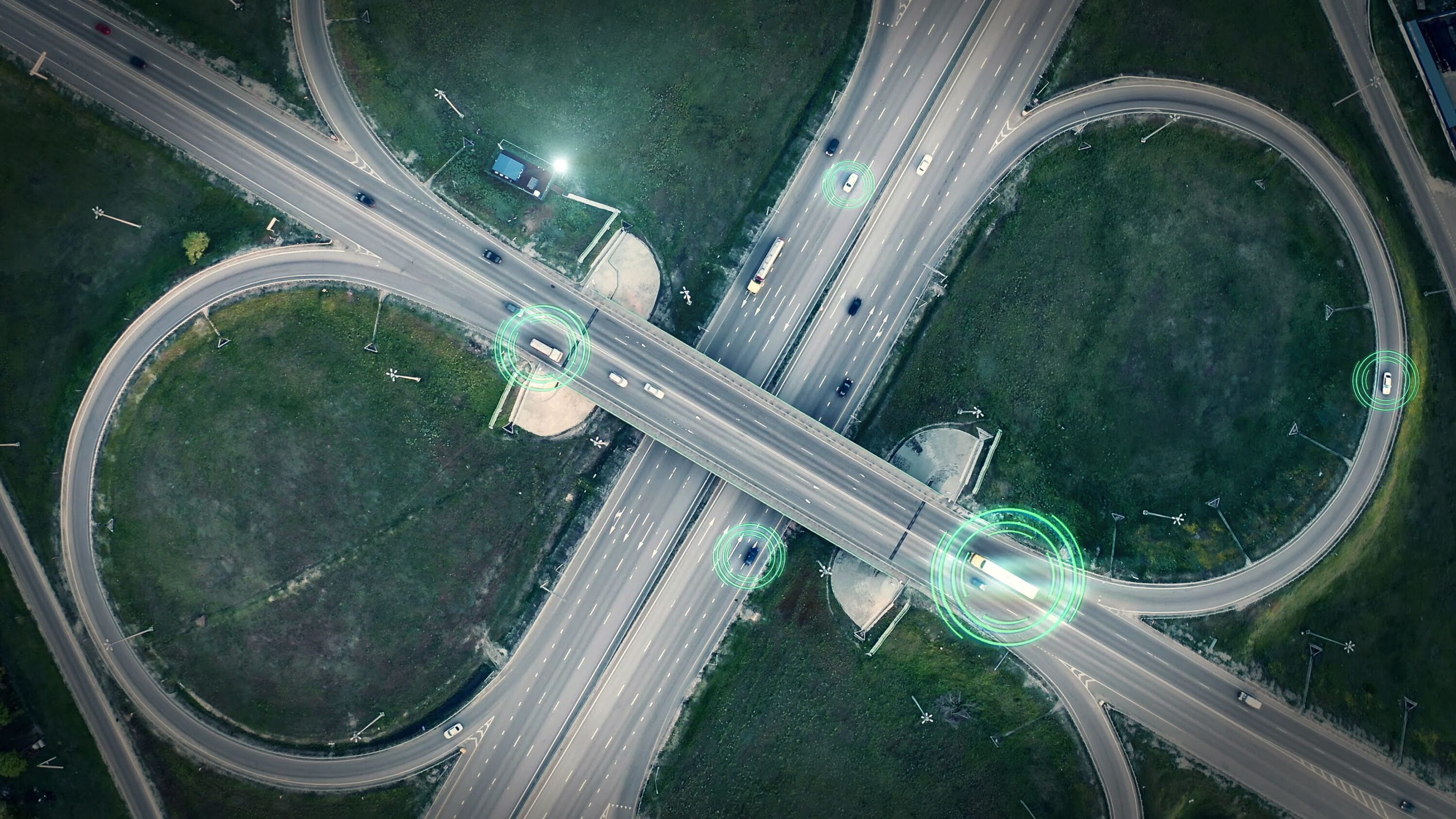 An aerial view of a cloverleaf highway interchange at dusk with digital green rings highlighting several vehicles on the road.