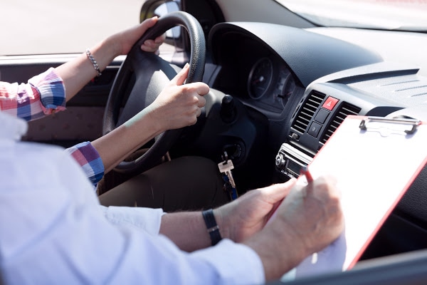 View from inside a car with a driver holding the steering wheel with 2 hands and the passenger writing on a clipboard.