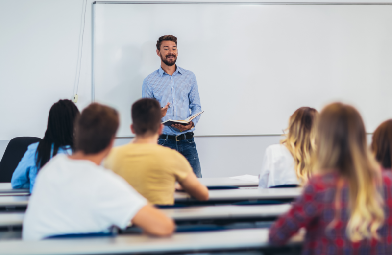 A classroom setting with multiple students sitting at desks and a male teacher standing at the front of the classroom.