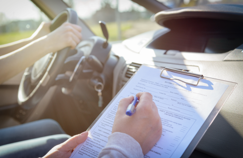 A close-up of an examiner holding a clipboard and pen in the passenger seat while a student drives the car.