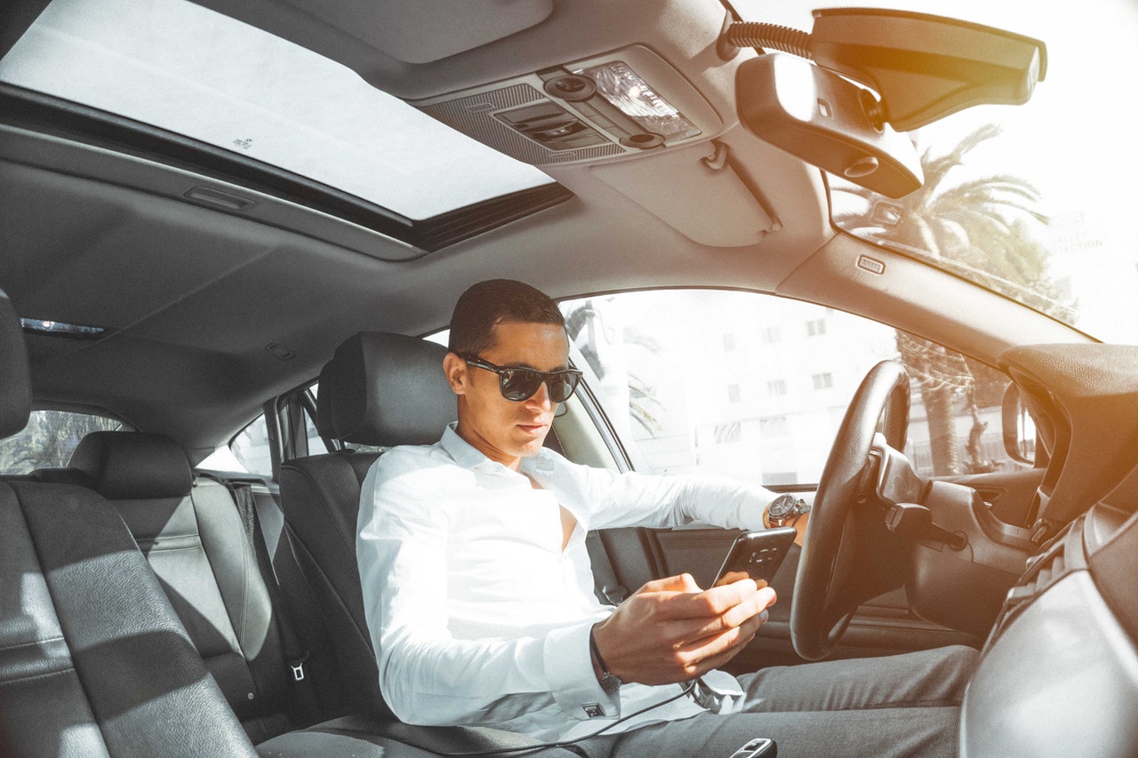 A man wearing sunglasses and a white button-down shirt looking down at a smartphone in his hand while sitting in the driver's seat of a car.