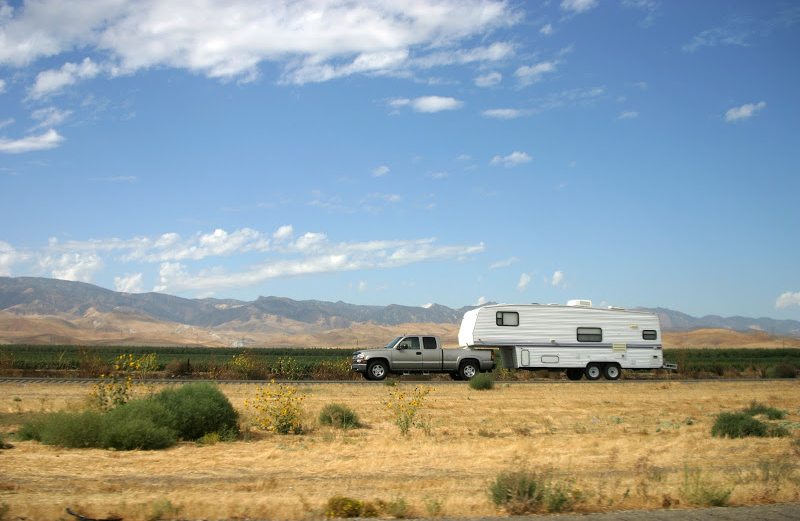 A white pickup truck towing a large white recreational travel trailer through a flat, grassy desert landscape under a blue sky with light clouds.