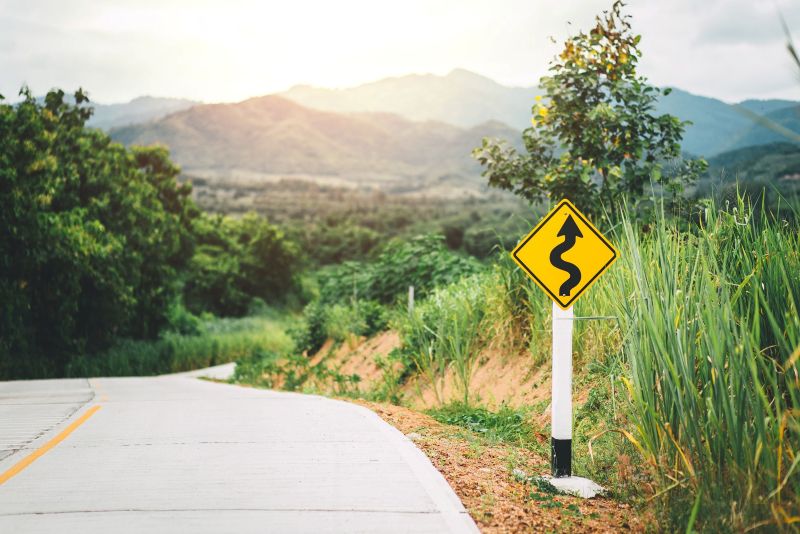A yellow diamond-shaped road sign with a black squiggly arrow indicating a winding road stands beside a paved mountain road, with rolling green hills and mountains fading into a bright, hazy sky in the distance.