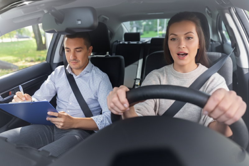A young woman with a wide-eyed, focused expression grips the steering wheel of a car, while a male driving examiner in the passenger seat intently writes notes on a blue clipboard.