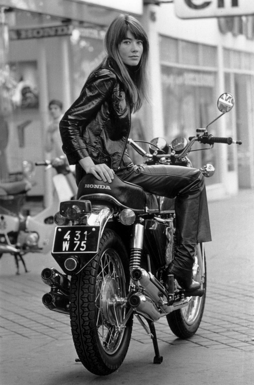 A black-and-white photo of a woman in leather clothing sitting on a Honda motorcycle parked on a sidewalk.