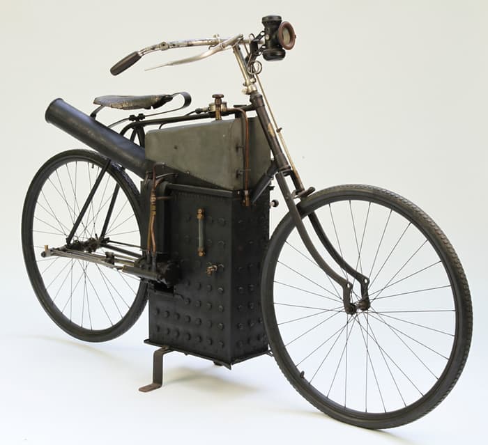 A studio shot of a very early motorized bicycle featuring a large, boxy black engine and a high-mounted exhaust pipe.