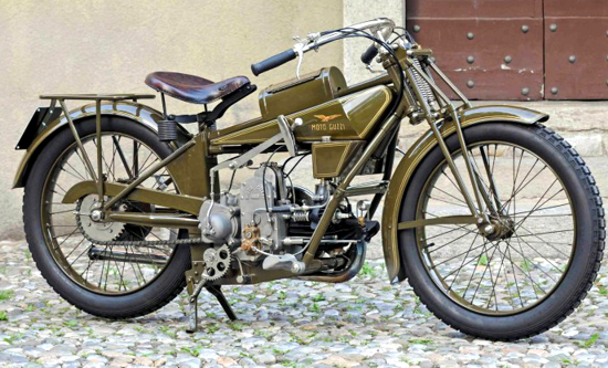 A side view of a vintage olive-green Moto Guzzi motorcycle parked on a cobblestone surface.
