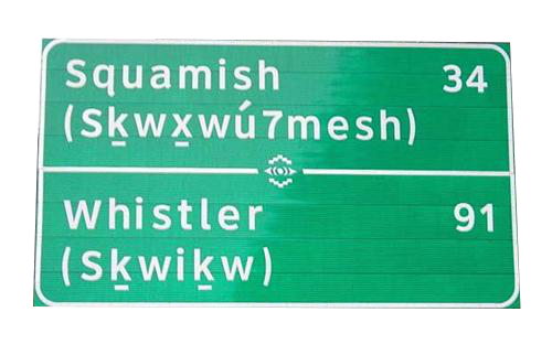 A highway sign displaying both English and traditional First Nations language names for a community.