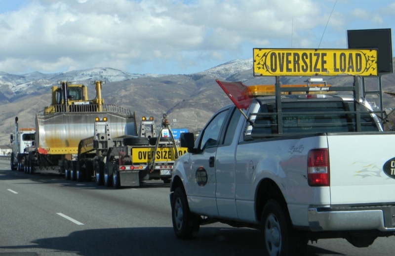A white pilot car with an "OVERSIZE LOAD" sign follows a semi-truck hauling a massive yellow bulldozer on a highway, with snow-capped mountains in the background.