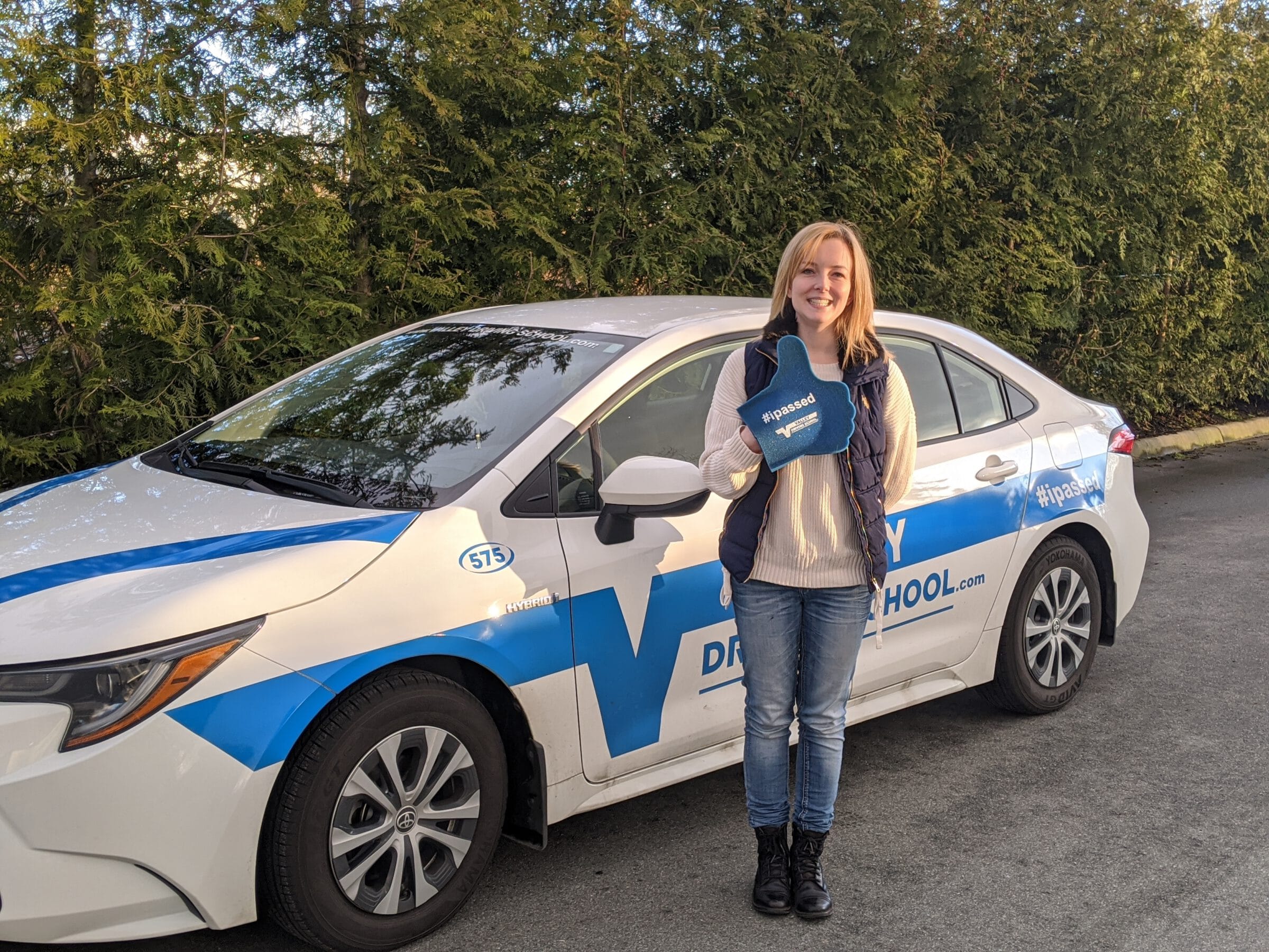 a young woman standing in front of a Valley Driving School car. She is smiling and holding a sign that says "I passed".