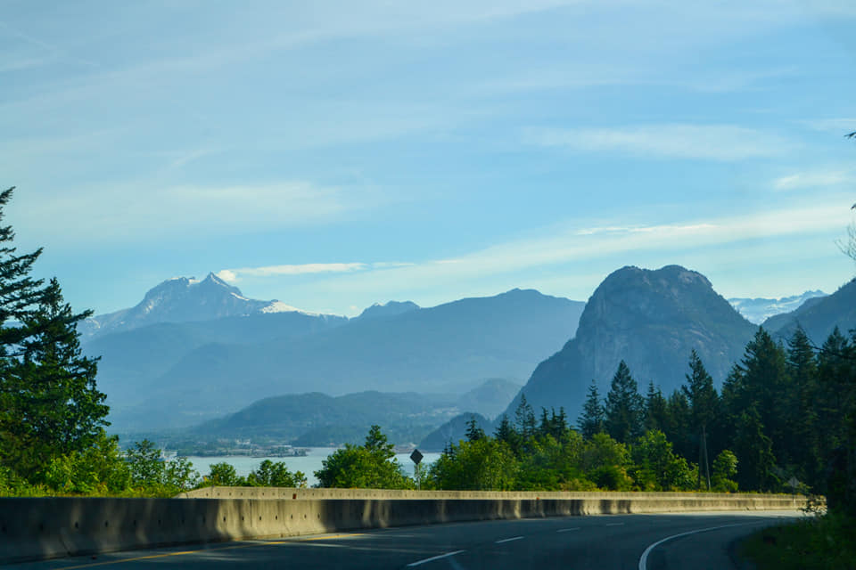 A view from a curved highway overlooking a body of water with large, hazy mountains and evergreen trees in the distance. Sea-to-Sky Highway, BC.