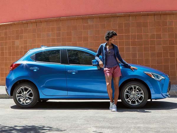 A woman leaning against a bright blue hatchback car parked in front of a brown tiled wall.