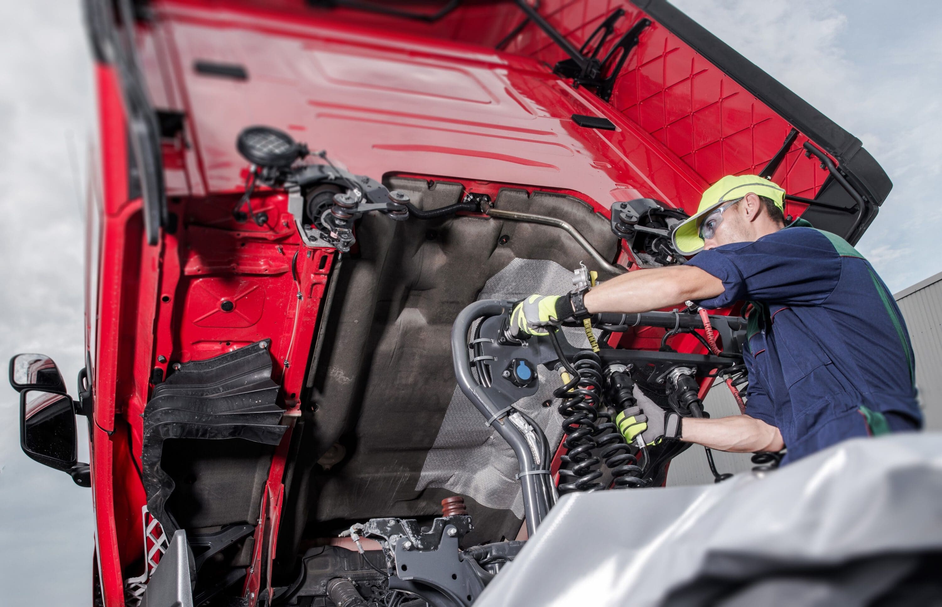 A mechanic in a yellow safety hat and gloves working on the underside of a tilted red semi-truck cab.
