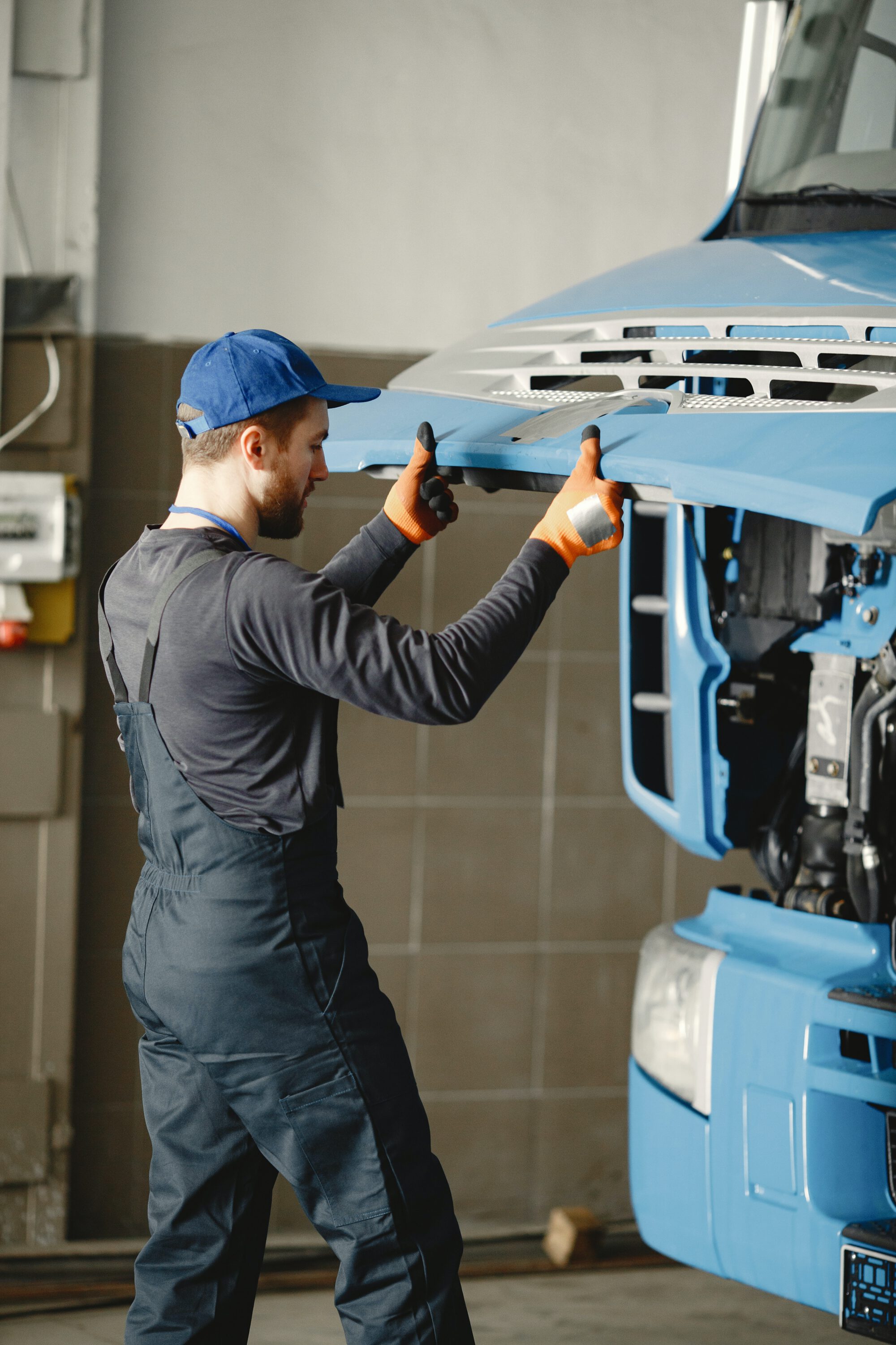 A mechanic in a blue uniform and a cap working on the engine of a blue semi-truck with the hood tilted open.
