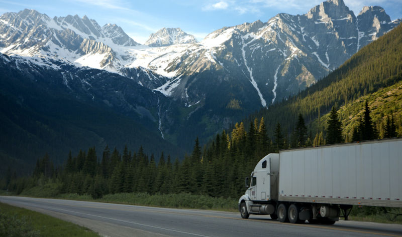 A white semi-truck with a trailer driving on a highway through a mountain range with snow-capped peaks.
