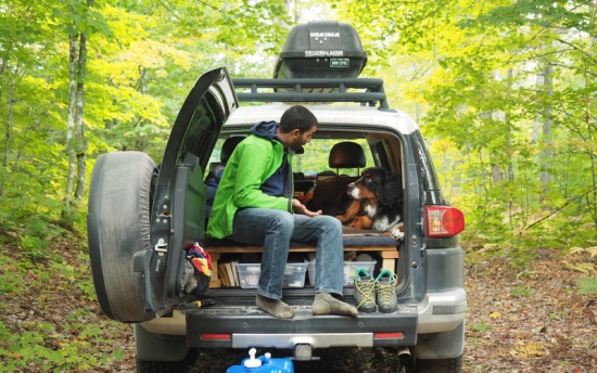 A man sitting on the edge of the open trunk of a dark green SUV parked in a forest; a medium-sized dog sits inside the vehicle beside him, and camping gear is visible on the roof rack.
