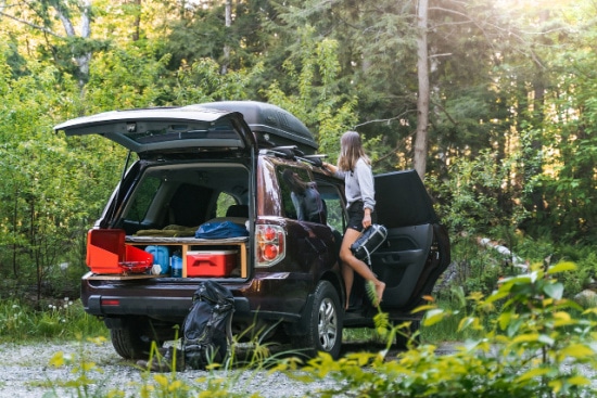 A woman stands by a dark SUV outfitted for camping with a rooftop cargo box and a custom wooden storage deck in the trunk containing a cooler and supplies, all parked in a lush, sun-dappled forest.