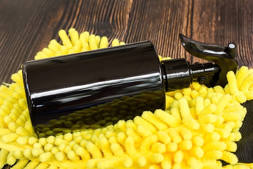 A black spray bottle resting on a yellow microfiber cleaning mitt on a dark wooden surface.