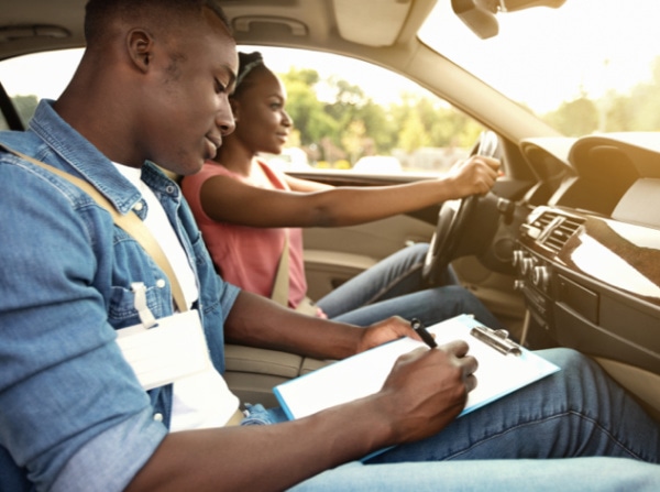 2 people in a car. a woman is driving and holding the steering wheel and a man is in the passenger seat, marking a clipboard.
