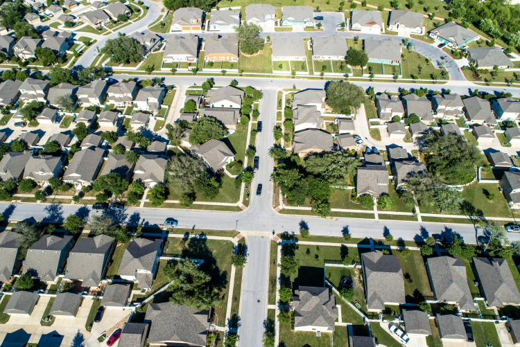 An aerial, bird’s-eye view of a quiet suburban neighborhood featuring rows of houses with grey roofs, manicured lawns, and paved streets forming a "T" intersection under bright sunlight.