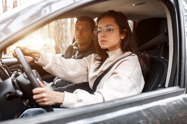 a young woman wearing glasses is behind the wheel of a car with a man sitting in the passenger seat, watching her drive.