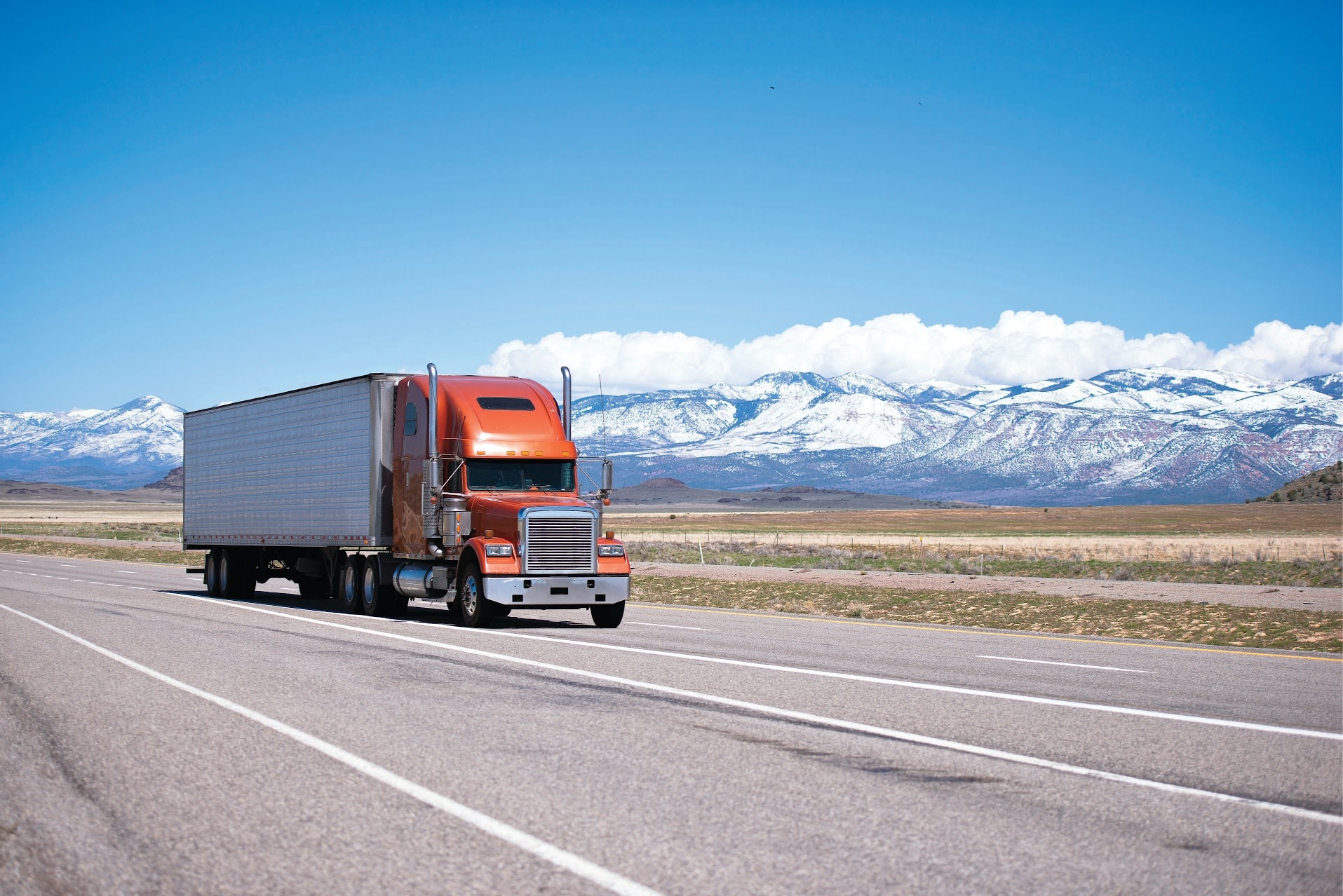 orange semi truck with white trailer driving down a road with snow capped mountains in the background.