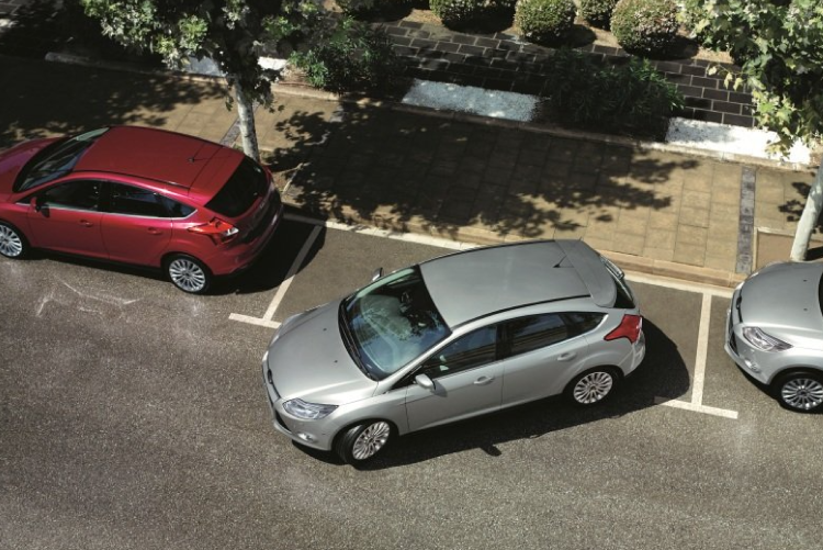 A silver car parallel parking into a tight spot on a city street between a red car and a grey car.