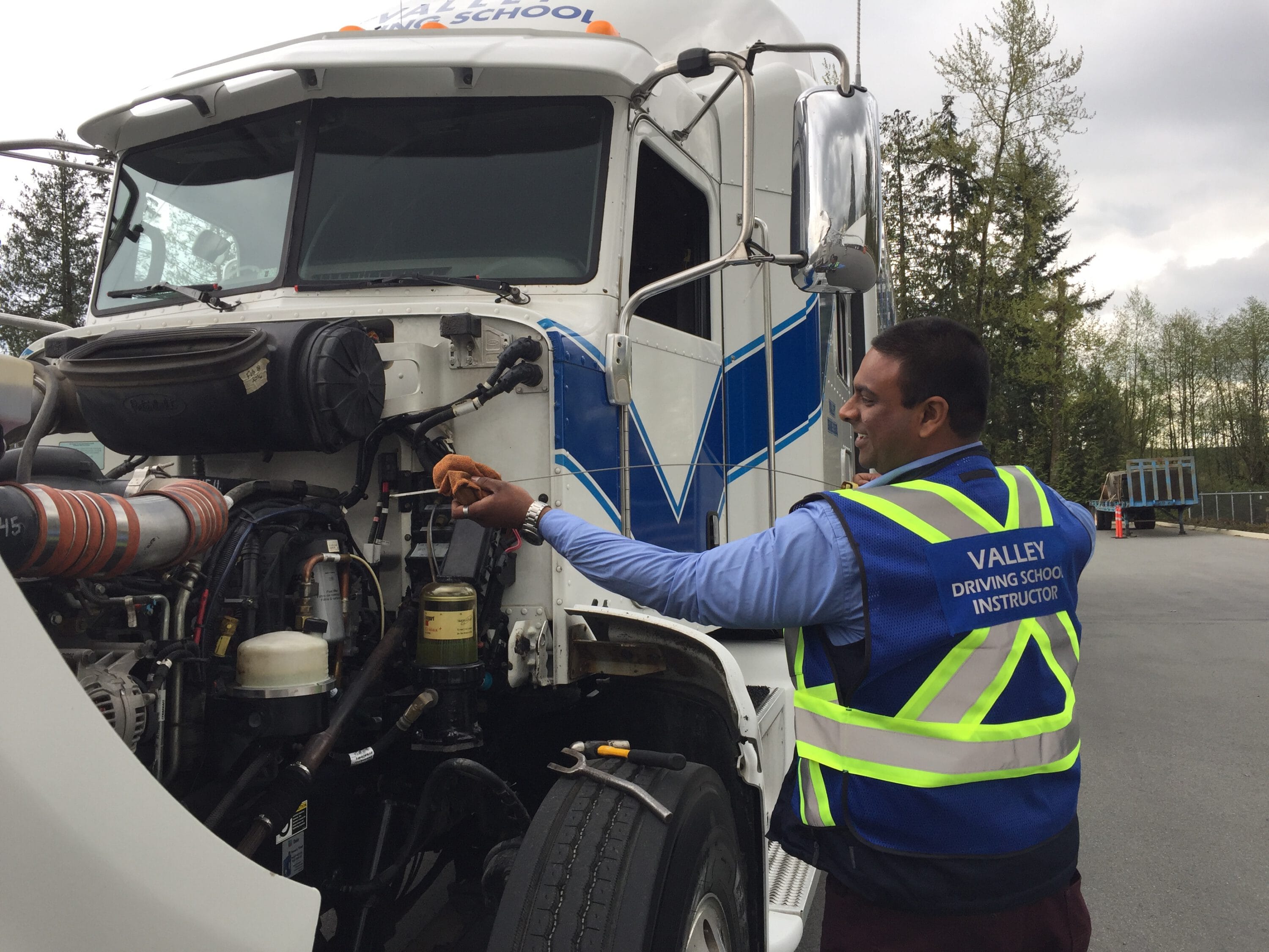 A man wearing a blue safety vest has the hood open on the semi truck and is checking the oil dipstick