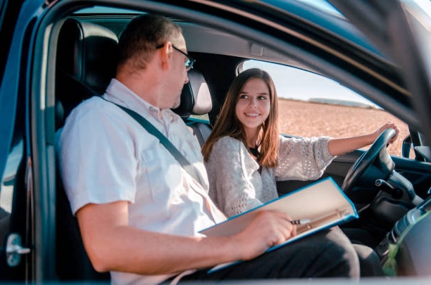 A young woman behind the wheel of a car during a driving lesson, with a male instructor in the passenger seat watching her drive.
