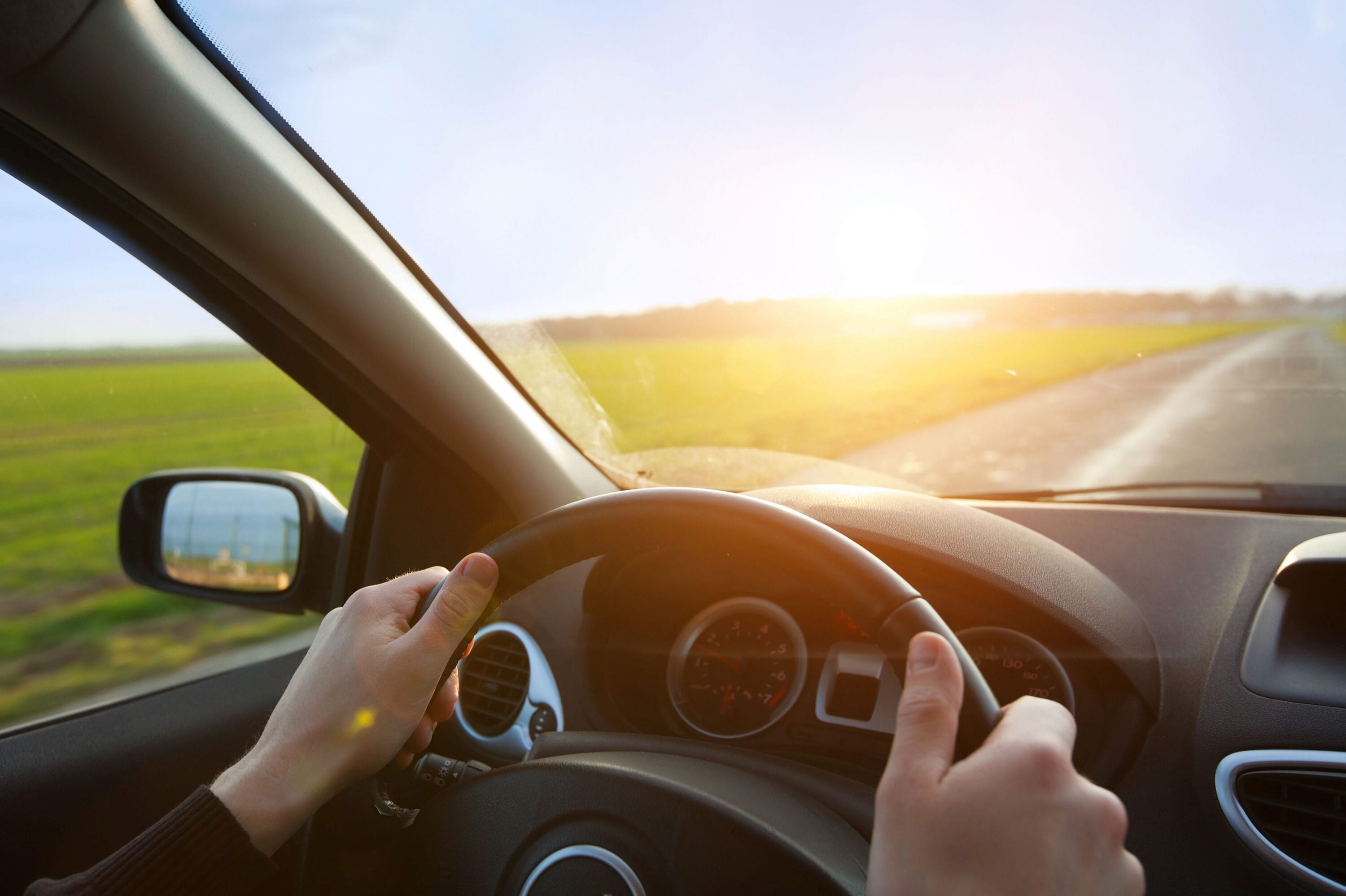 hands on the steering wheel of a car with the sunsetting in the distance.