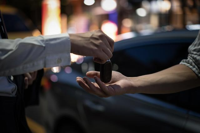 one person handing a car key to another person, while standing beside a dark coloured car.