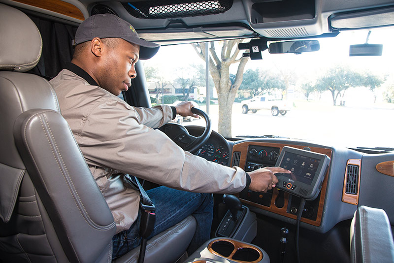 A truck driver in a tan jacket and baseball cap sitting in the driver's seat and operating an electronic logging device mounted on the dashboard.