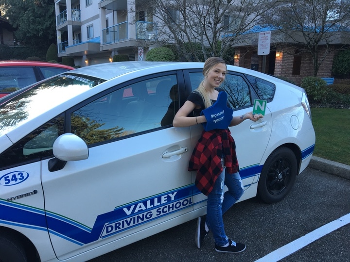 A student smiling and holding an I Passed sign while standing next to a Valley Driving School car.
