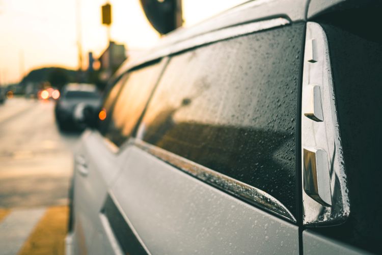 A close-up view of a white car's side windows covered in water droplets, with a blurred street scene and warm sunset light reflecting in the background.
