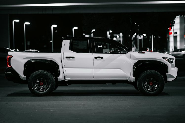 A high-contrast side profile of a white pickup truck with black wheels, fender flares, and roof, featuring small red accents on the wheel centers, parked in an outdoor lot at night under streetlights.