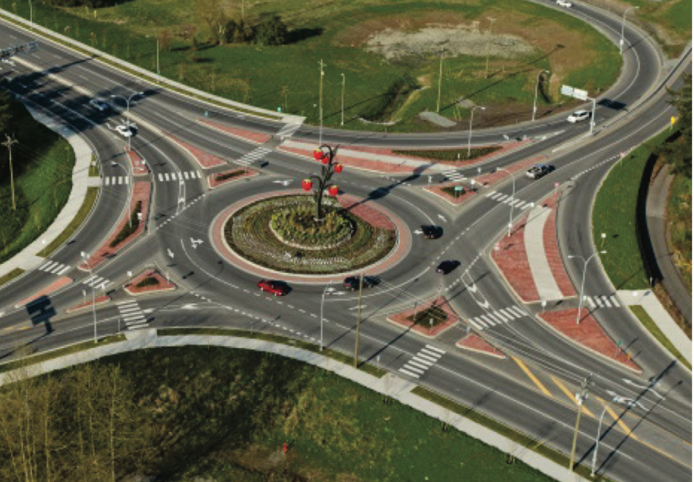 Multi lane roundabout in Abbotsford with cars driving. There is a raspberry statue in the grassy area in the middle of the roundabout.