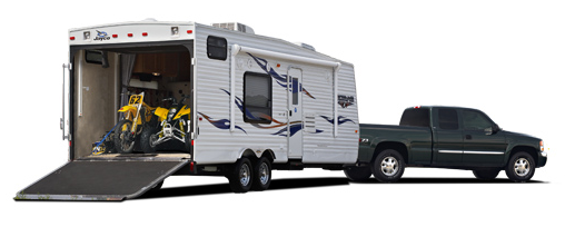 A dark green GMC pickup truck towing a white Jayco toy hauler travel trailer with the rear ramp open, revealing a yellow dirt bike and ATV inside.