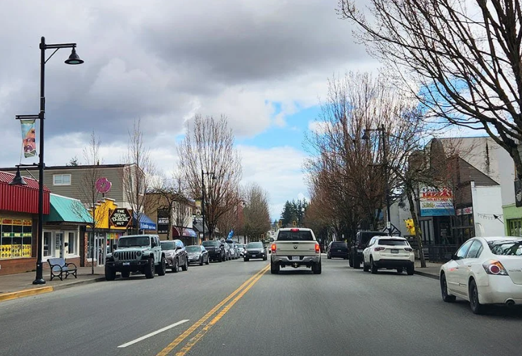 Downtown Aldergrove road with a pick up truck and car driving on the road.  There is local businesses and parked cars lining the roadway.