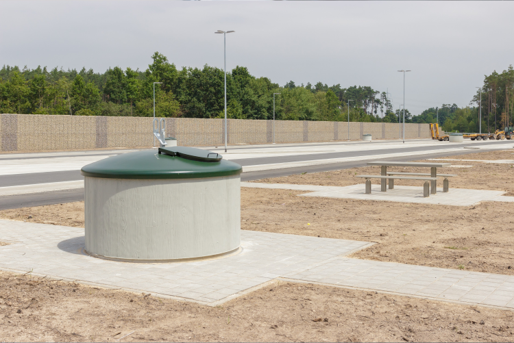 A large, cylindrical concrete waste bin with a dark green lid sits on a paved walkway at a highway rest area, with a picnic table and a long noise barrier wall in the background.