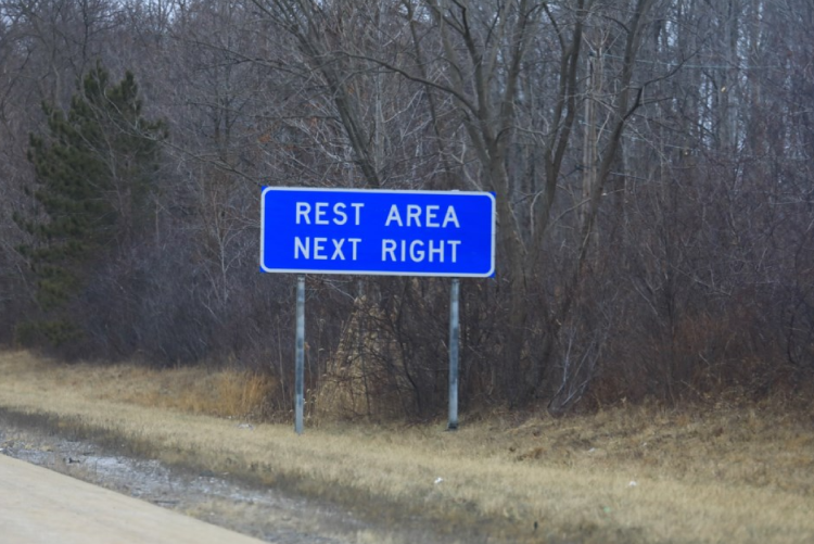A blue rectangular highway sign that reads "REST AREA NEXT RIGHT" in white capital letters, standing on a grassy roadside in front of a backdrop of bare trees.