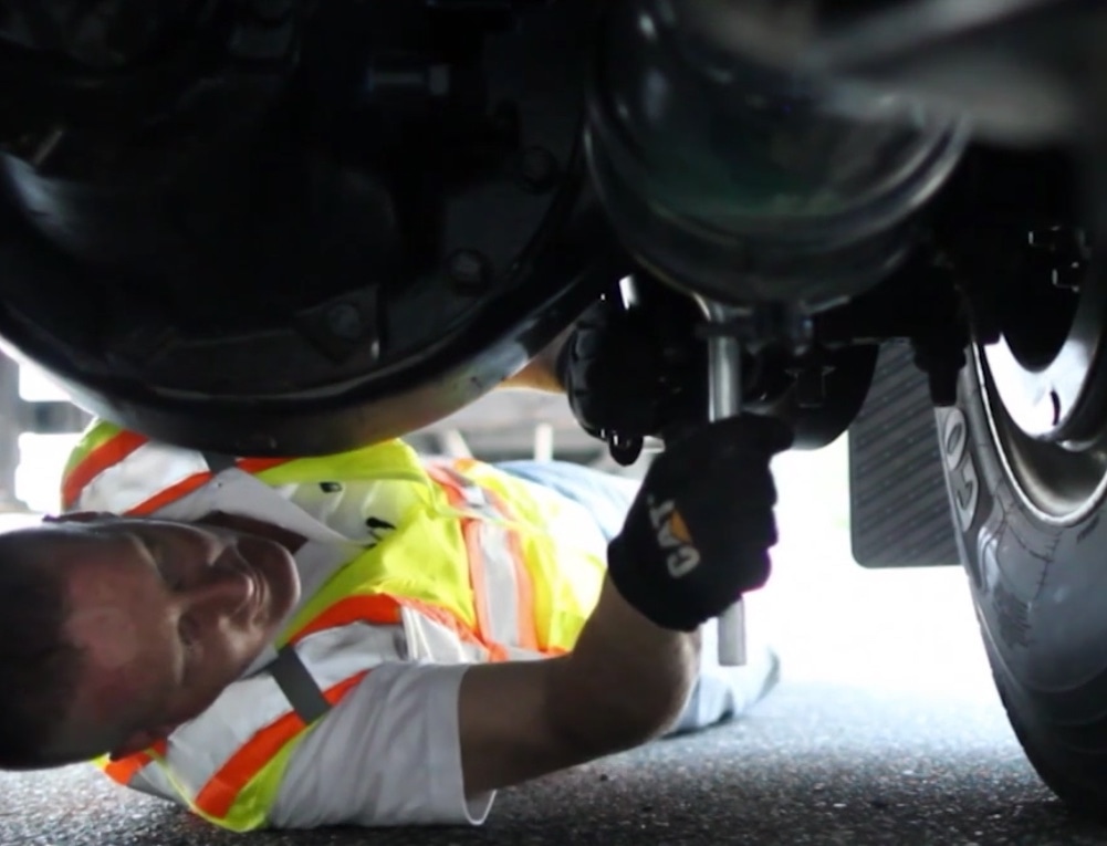 Man underneath a semi truck, wearing a yellow safety vest while checking the air brake system.