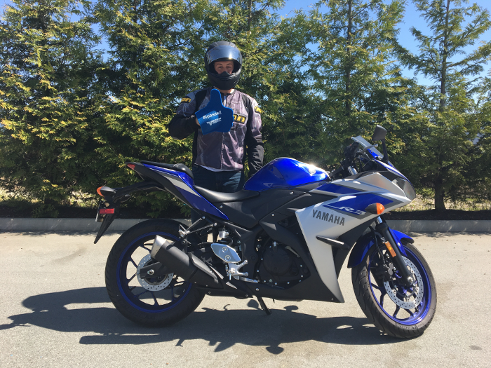 a motorcycle rider in full gear and helmet, standing behind a blue and silver sports bike.