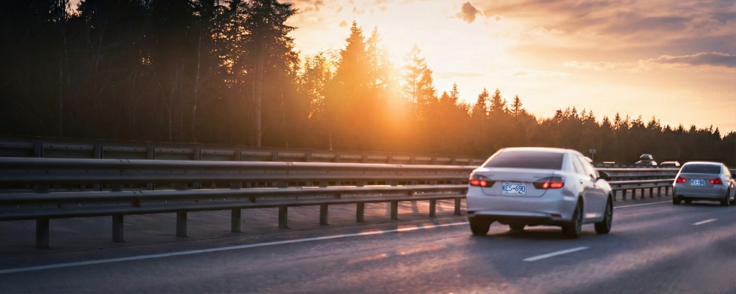 Sunset on a BC highway. There is a white car in the inside lane and another in front of it.