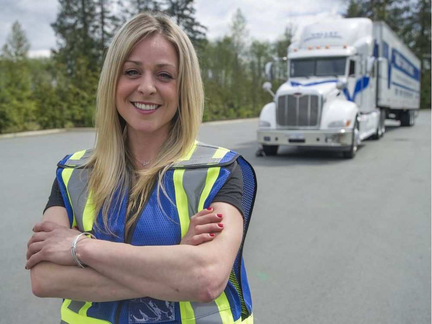 A smiling professional female driver in a blue safety vest standing with arms crossed in front of a Valley Driving School semi-truck.