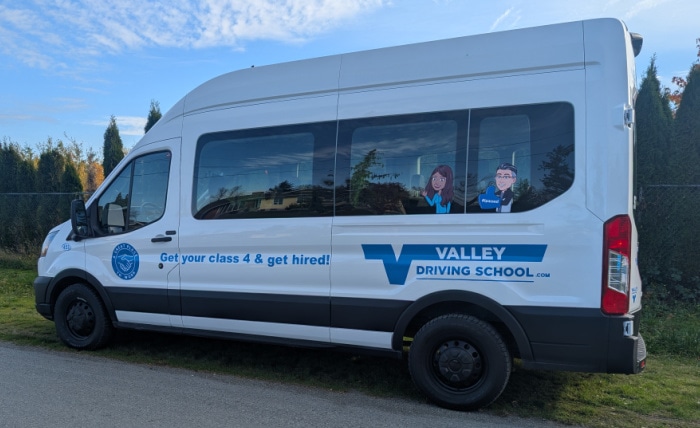 White Valley Driving School Ford Transit van with a decal reading "Get your class 4 & get hired!" parked on a gravel roadside.