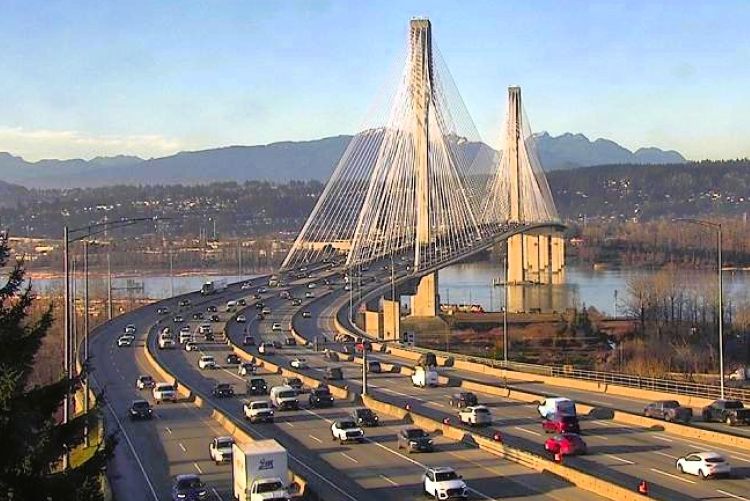 A high-angle view of the Port Mann Bridge with heavy traffic flowing across its multiple lanes, spanning a wide river with mountains visible in the distance.