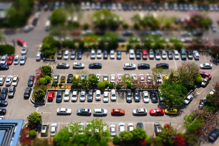 A high-angle, tilt-shift photograph of a busy outdoor parking lot filled with rows of colorful cars and interspersed with green trees and landscaping.