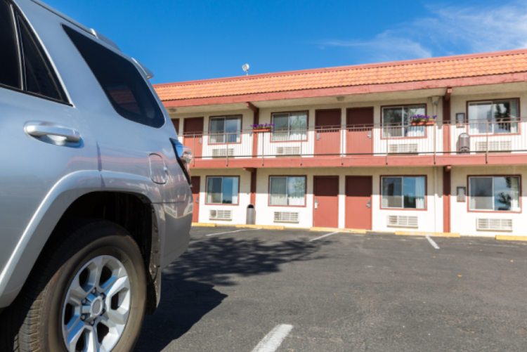 A silver SUV parked in an asphalt lot in front of a two-story motel building with red doors and white-trimmed windows under a clear blue sky.