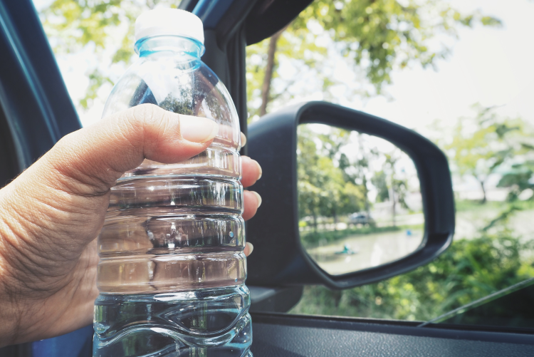 A hand holding a clear plastic water bottle inside a car, with the side-view mirror and lush green trees visible through the window in the background.